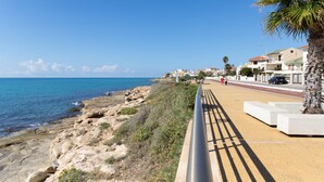 A coastal walkway with a rocky shoreline, a palm tree, and buildings in the background. - Apartment (Marina di Ragusa)