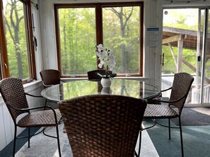A room with a round glass table and wicker chairs, a potted plant, and a view of trees through large windows.