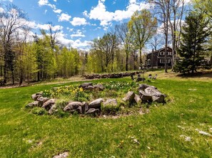 A landscaped garden with a stone circle, a wooden cabin, and a forested area.