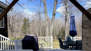 Ein Whirlpool mit Grill und Sonnenschirm, umgeben von einer Holzterrasse und weißem Geländer, mit Blick auf einen bewaldeten Bereich.