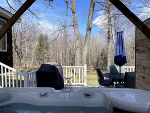 Ein Whirlpool mit Grill und Sonnenschirm, umgeben von einer Holzterrasse und weißem Geländer, mit Blick auf einen bewaldeten Bereich.