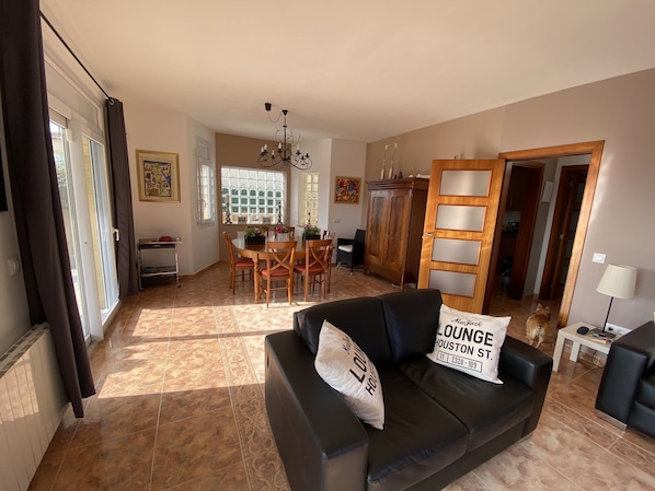 A spacious living area with a black leather sofa, a dining table with chairs, a chandelier, and a large wooden cabinet.