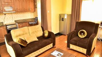 Una sala de estar con sofá y sillón, un área de cocina con armarios y estufa, y un refrigerador.