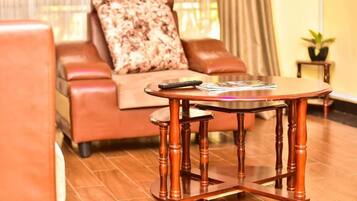 A brown leather sofa with a patterned pillow, a round wooden table with a remote control and magazines on it, and a small potted plant on a side table.