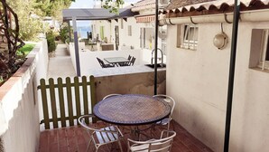 Um pátio com uma mesa redonda e duas cadeiras, uma área de estar coberta e uma vista para um prédio branco com telhado de azulejos.