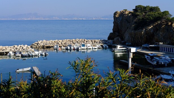 Uma marina com barcos atracados, um paredão rochoso e vista clara para o mar.