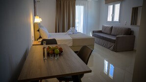 A hotel room with a bed, a sofa, a dining table with fruit and glasses, and a window with curtains.