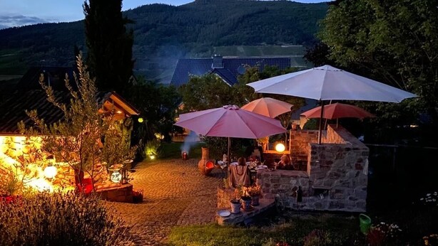 Un espace extérieur aménagé pour les repas, avec des tables en pierre et des parasols rouges, entouré d’une végétation luxuriante et d’un mur de pierre.