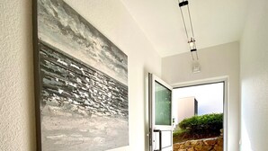 Un couloir orné d’un grand tableau abstrait au mur, une porte donnant sur l’extérieur et un mur de pierre visible à travers cette porte.