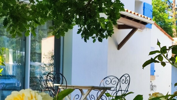 Ein Garten mit Tisch und Stühlen, ein weißes Gebäude mit blauen Fensterläden und eine gelbe Rose im Vordergrund.
