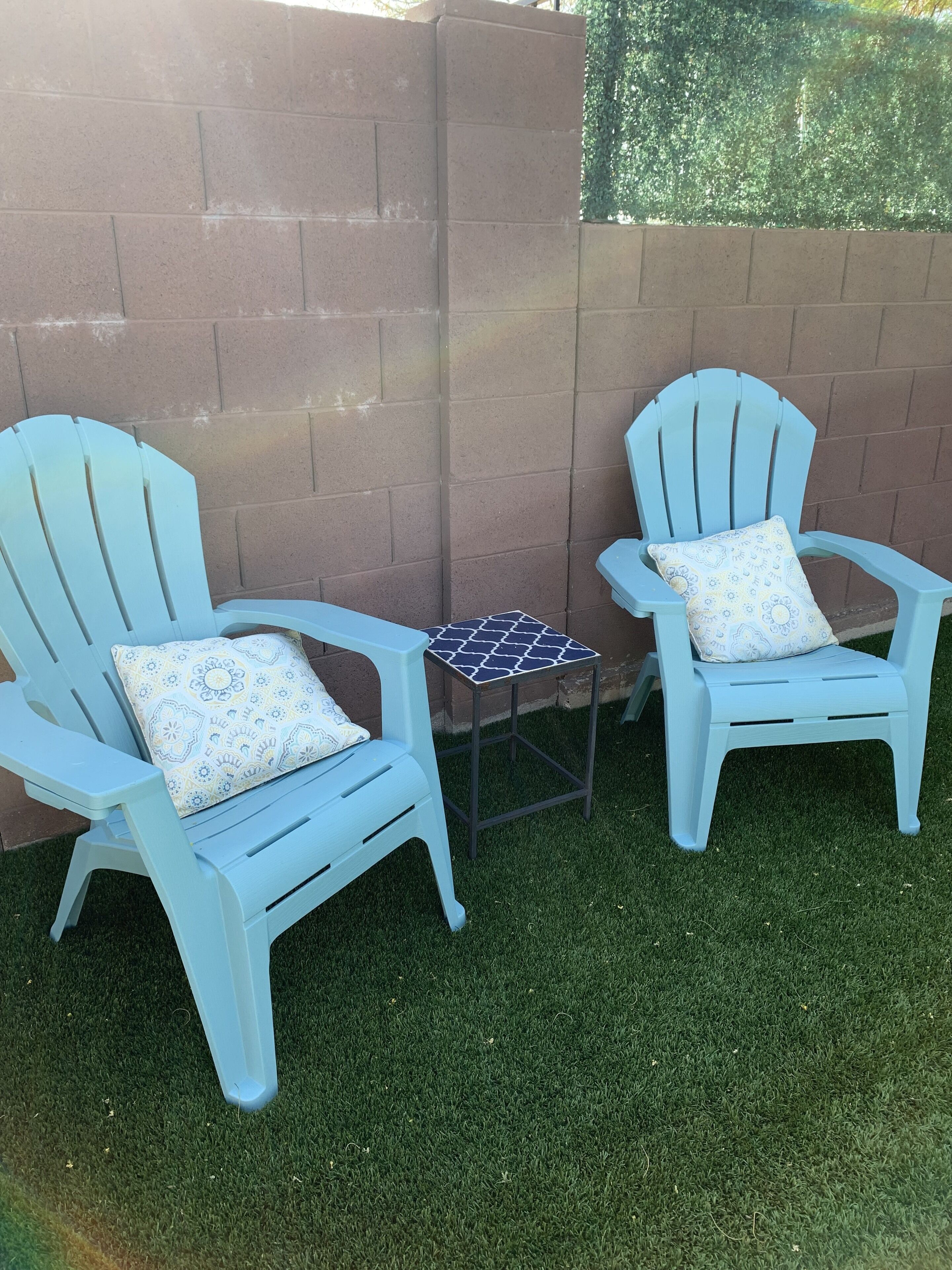 Twee lichtblauwe Adirondack-stoelen met gestreepte kussens, een klein vierkant tafeltje met een blauw-witte blad, en een bakstenen muur op de achtergrond.