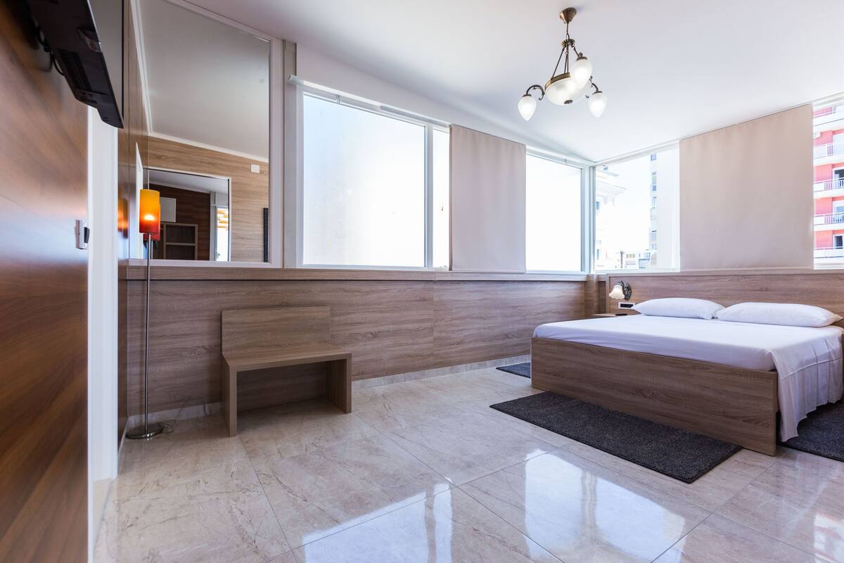 A modern hotel room with a large bed, a bench, a desk, and a large window offering a city view.
