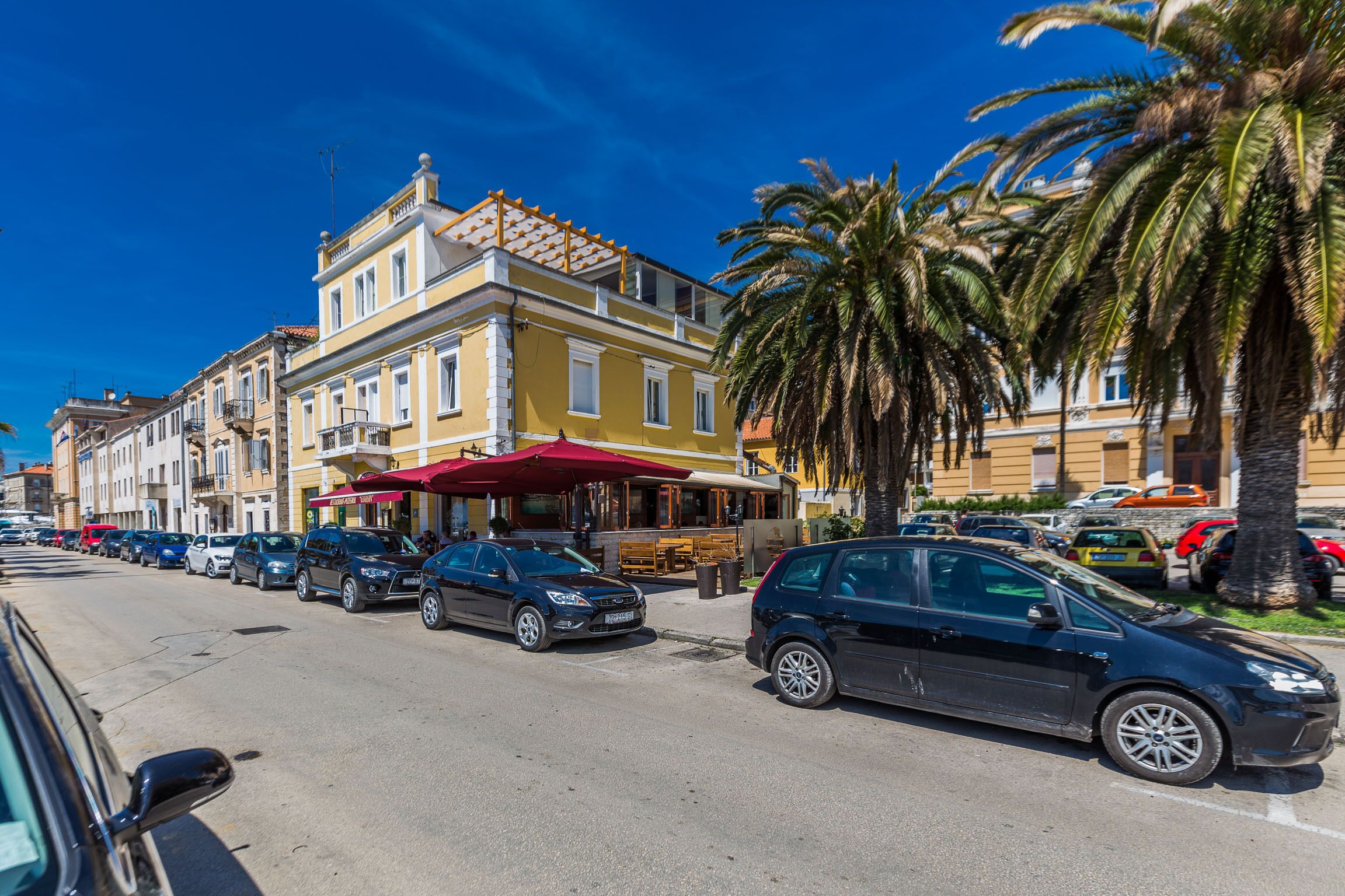 A street lined with parked cars and a row of buildings with balconies and a red awning.