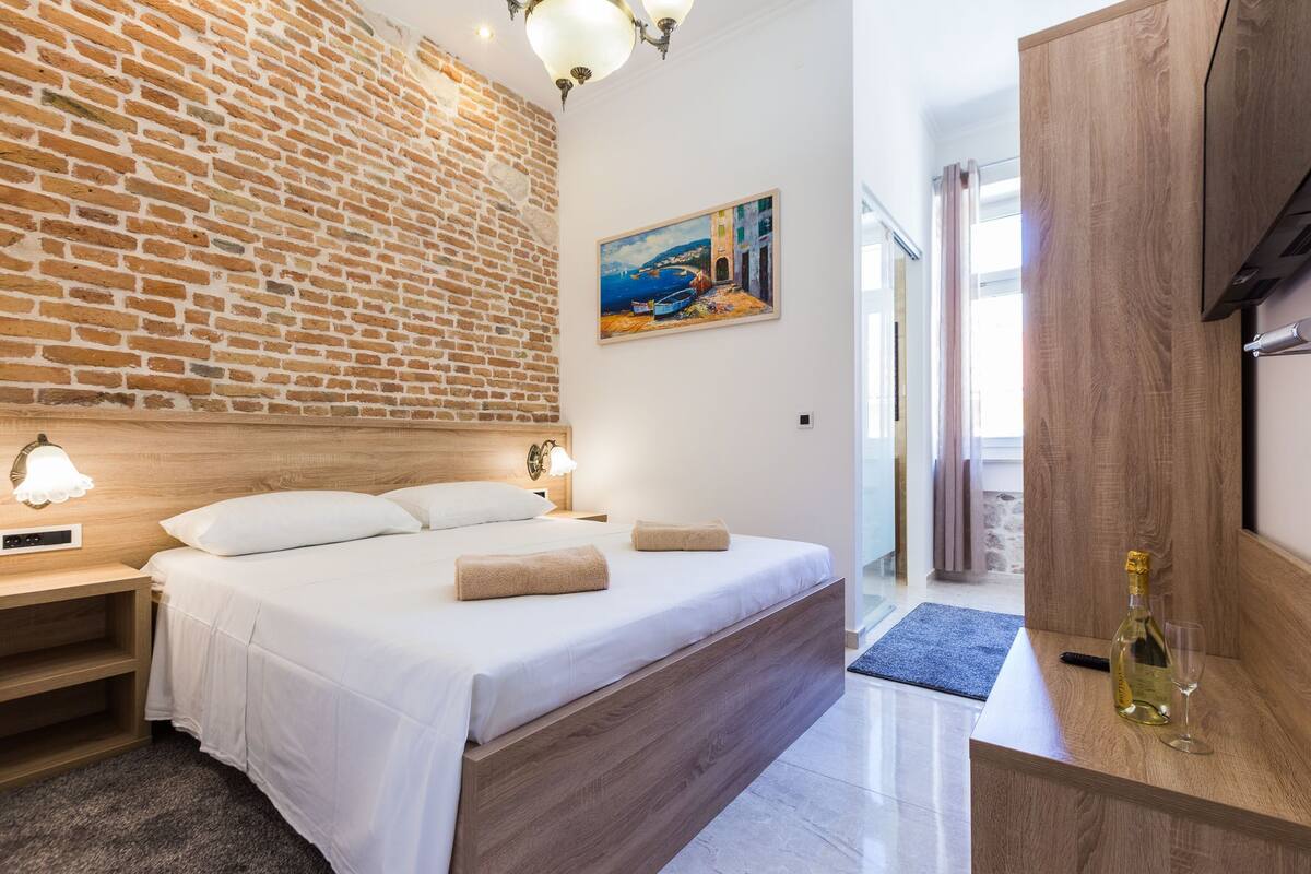 A modern hotel room with a brick wall, a painting of a coastal scene, a wooden bed, a nightstand with a lamp, and a minibar with a bottle and glass.
