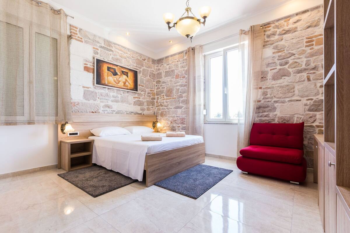 A bedroom with a stone wall, a bed with white bedding, a red chair, and a nightstand with a lamp.