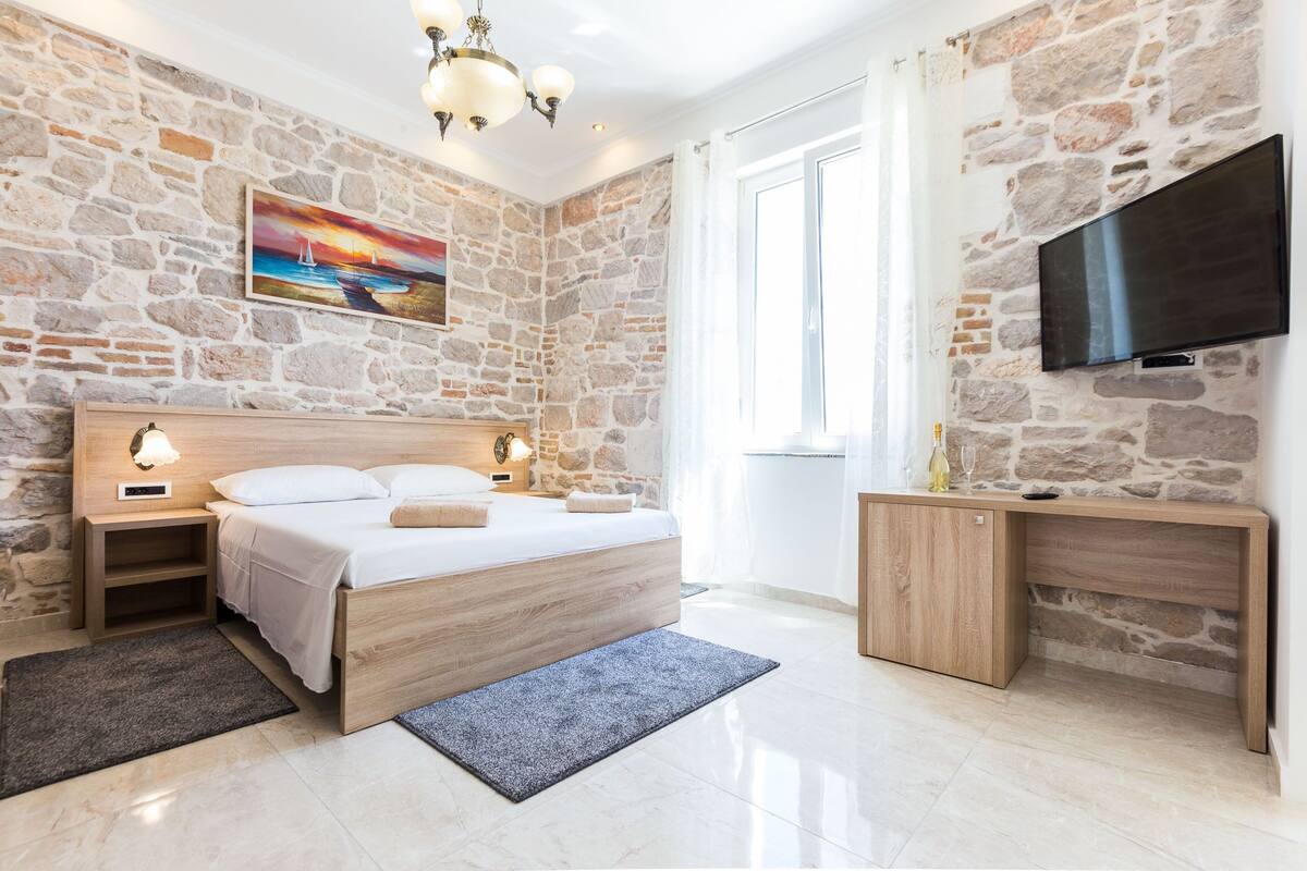 A bedroom with a stone wall, a bed, bedside tables, a TV, and a window.