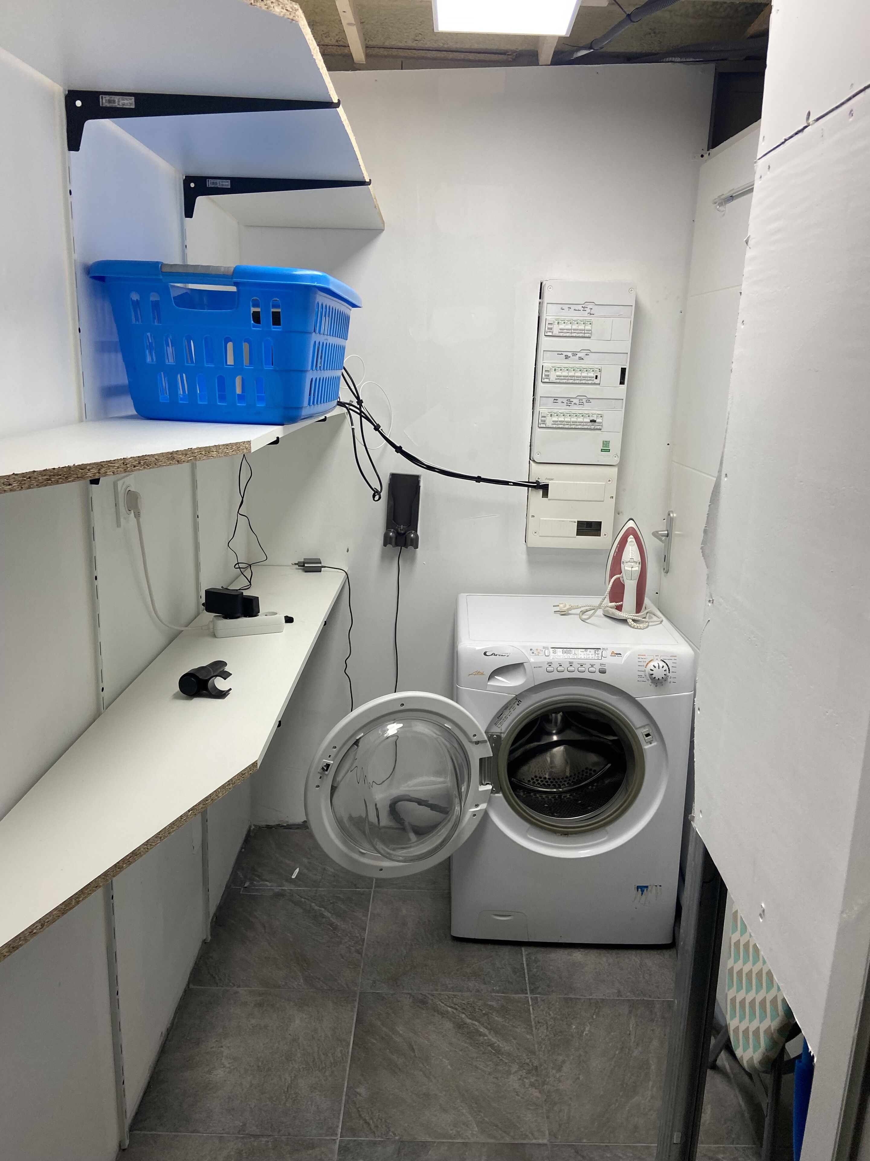 A compact laundry room with a washing machine, a blue laundry basket on a shelf, and electrical outlets.