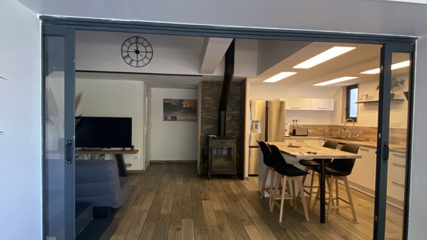 A modern kitchen with a built-in oven, a dining area with a wooden table and chairs, and a living room with a television and a fireplace.