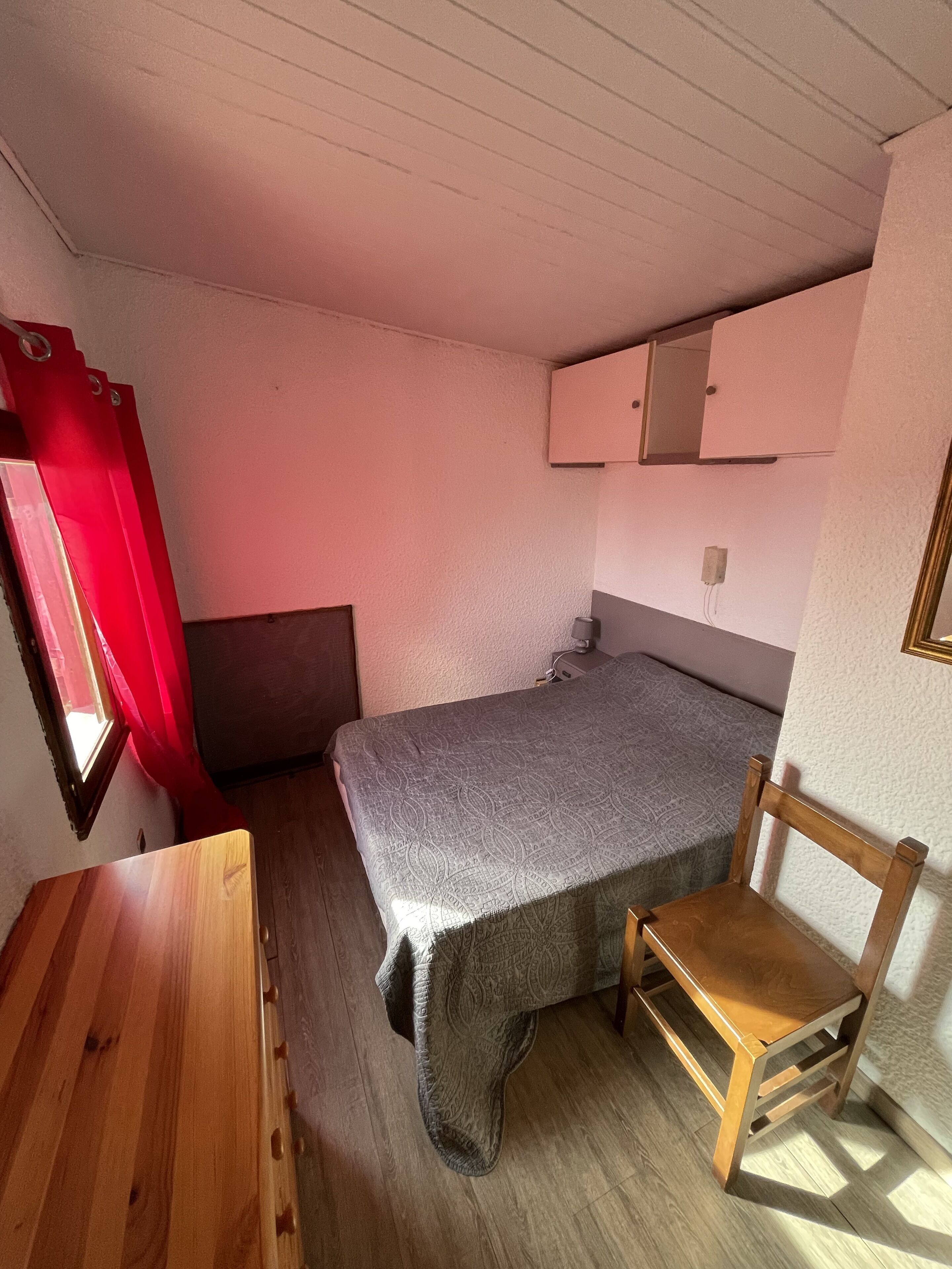 Ett litet, kompakt rum med en enkel säng, en trästol, ett litet bord och ett fönster med röda gardiner.