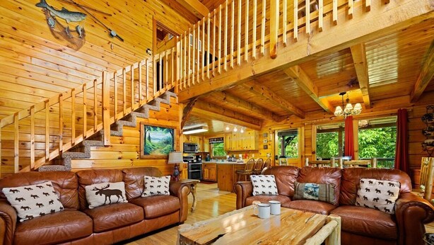 Ein Blockhaus-Interieur mit einer Holztreppe, Ledersofas mit Rentier-Muster Kissen, einem Holzes Esstisch und einer Küchenzeile.