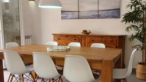 Een eethoek met een houten tafel, witte stoelen, een hanglamp, een decoratiemkast en een schilderij aan de muur.