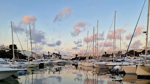 Een jachthaven met meerdere zeilboten afgemeerd, kalm water dat de boten en lucht weerspiegelt, en gebouwen op de achtergrond.
