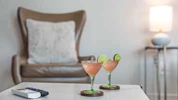 Um controle remoto, duas bebidas com limão em uma mesa, uma cadeira marrom e uma luminária ao fundo.