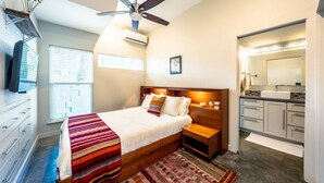 Una camera da letto con un letto, comodini, una ventola a soffitto, una finestra e un bagno con lavandino e specchio.