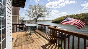 A wooden deck with a table and chairs, overlooking a river with an American flag. - Lakefront Family Retreat | Sleeps 10+ | Pet-Friendly | Swim & Fish Off the Dock (Sunrise Beach)