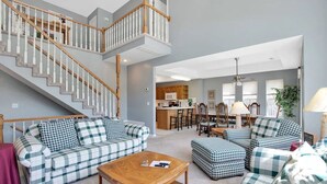 A spacious living area with a staircase, checkered sofas, a wooden coffee table, and a kitchen in the background. - Lakefront Family Retreat | Sleeps 10+ | Pet-Friendly | Swim & Fish Off the Dock (Sunrise Beach)