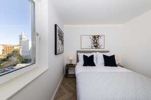 A hotel room with a large window, a bed with white bedding and black pillows, a nightstand with a lamp, and two framed artworks on the wall.