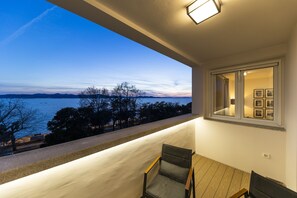 A balcony with a view of the sea and mountains at dusk. There is a table with chairs and a window with framed pictures.