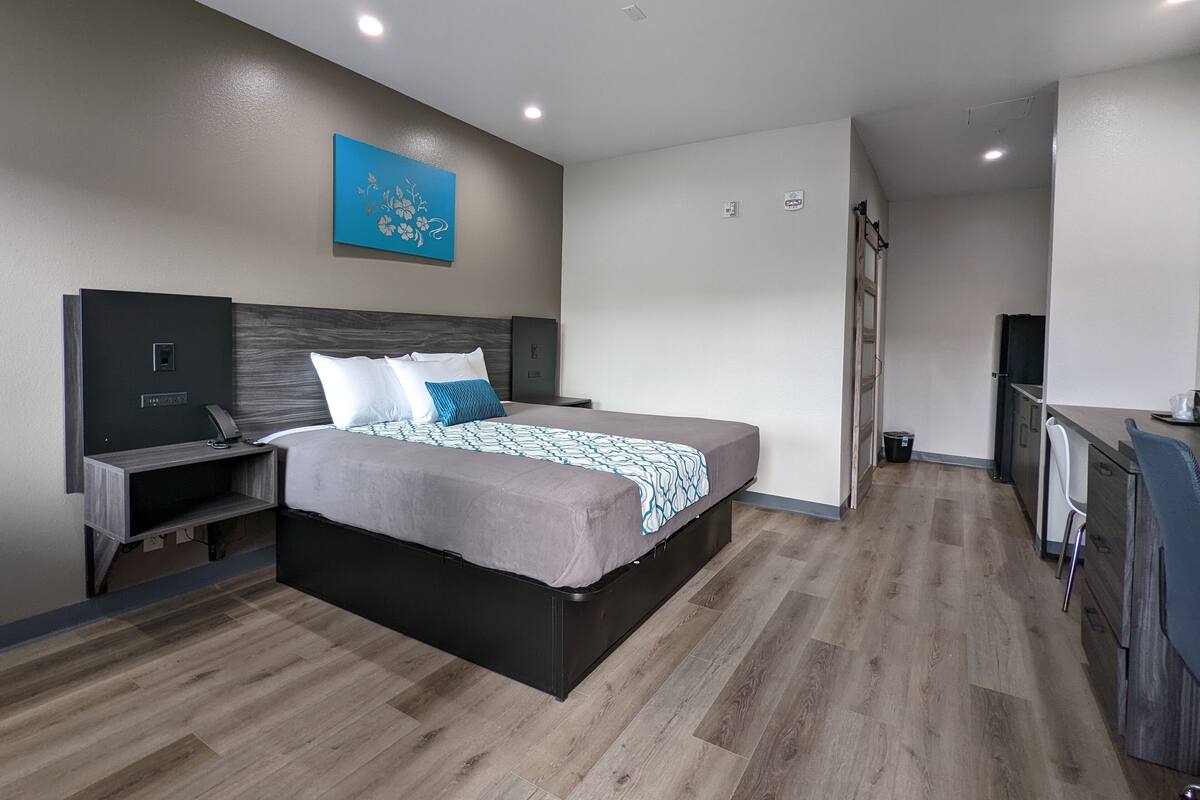 A modern hotel room with a large bed, a desk with a chair, a small TV, and a kitchenette area.