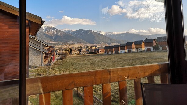 Une vue depuis une fenêtre donnant sur une zone rurale, avec des bâtiments en bois et des montagnes en arrière-plan.