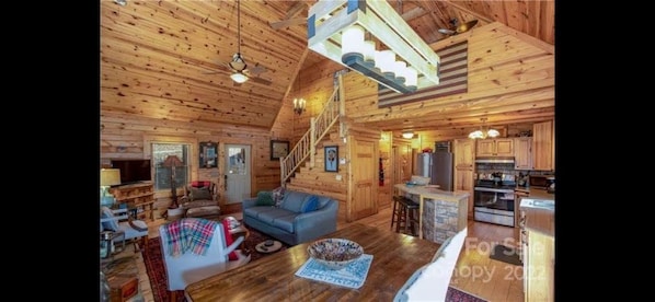 A wooden cabin interior with a living room, kitchen, and a staircase leading to an upper level.