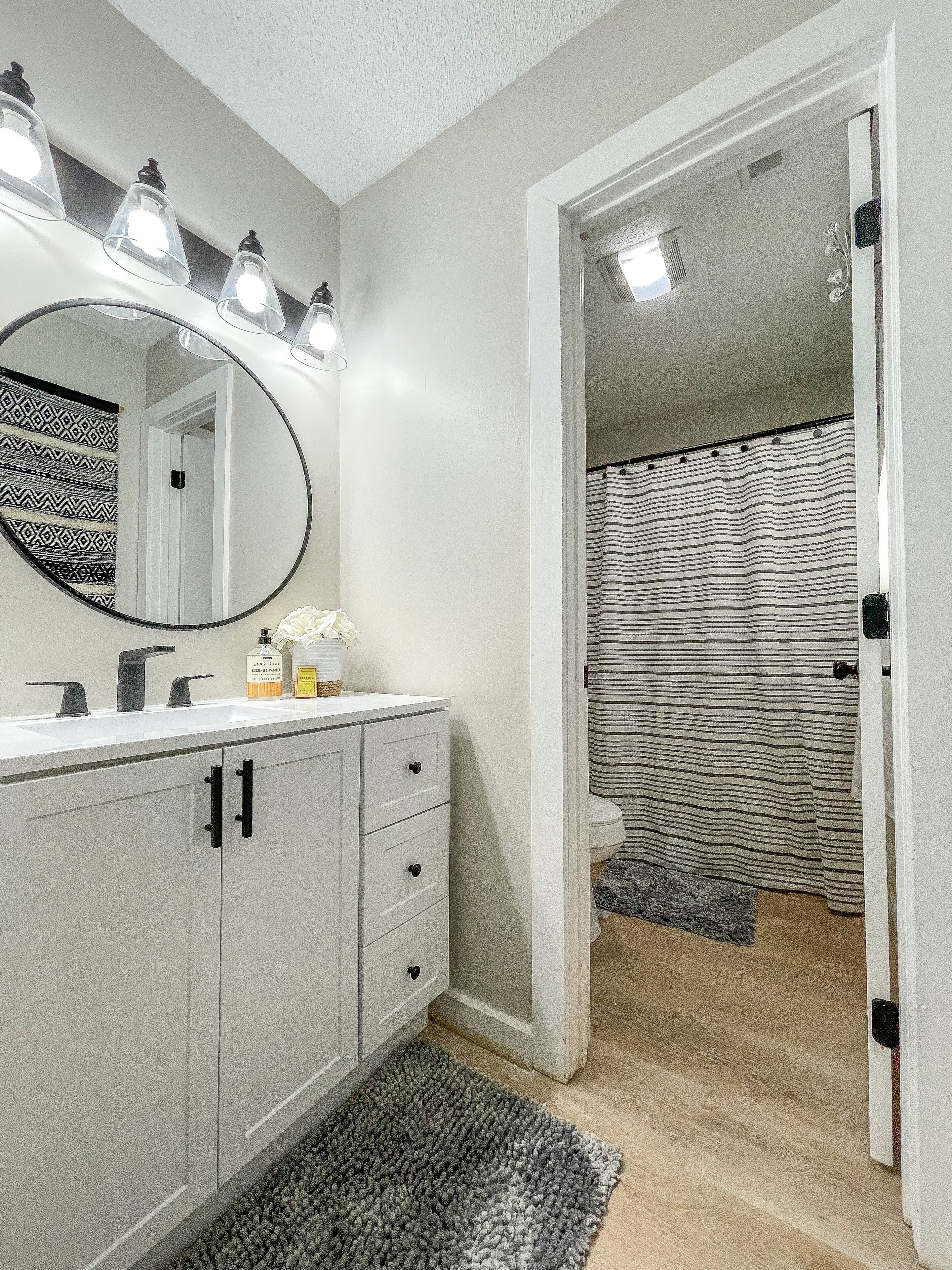 A bathroom with a white vanity, a round mirror with lights, a striped shower curtain, and a gray rug.