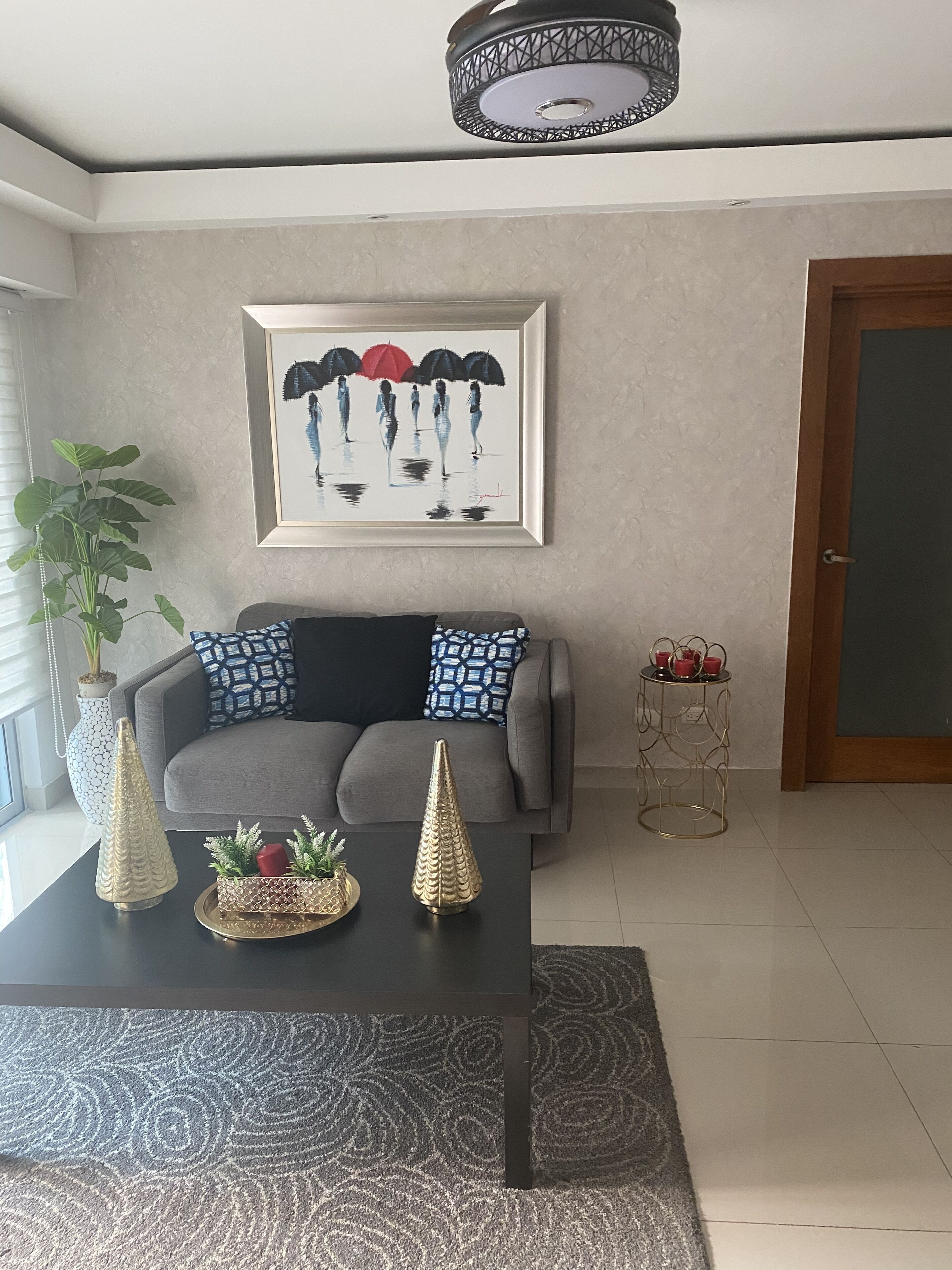 A living room with a grey sofa, a black coffee table, a framed artwork above the sofa, a potted plant, and a small side table with red cups.