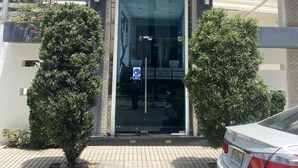 Entrada de un edificio moderno con puerta de cristal, palmeras y un coche blanco estacionado cerca.