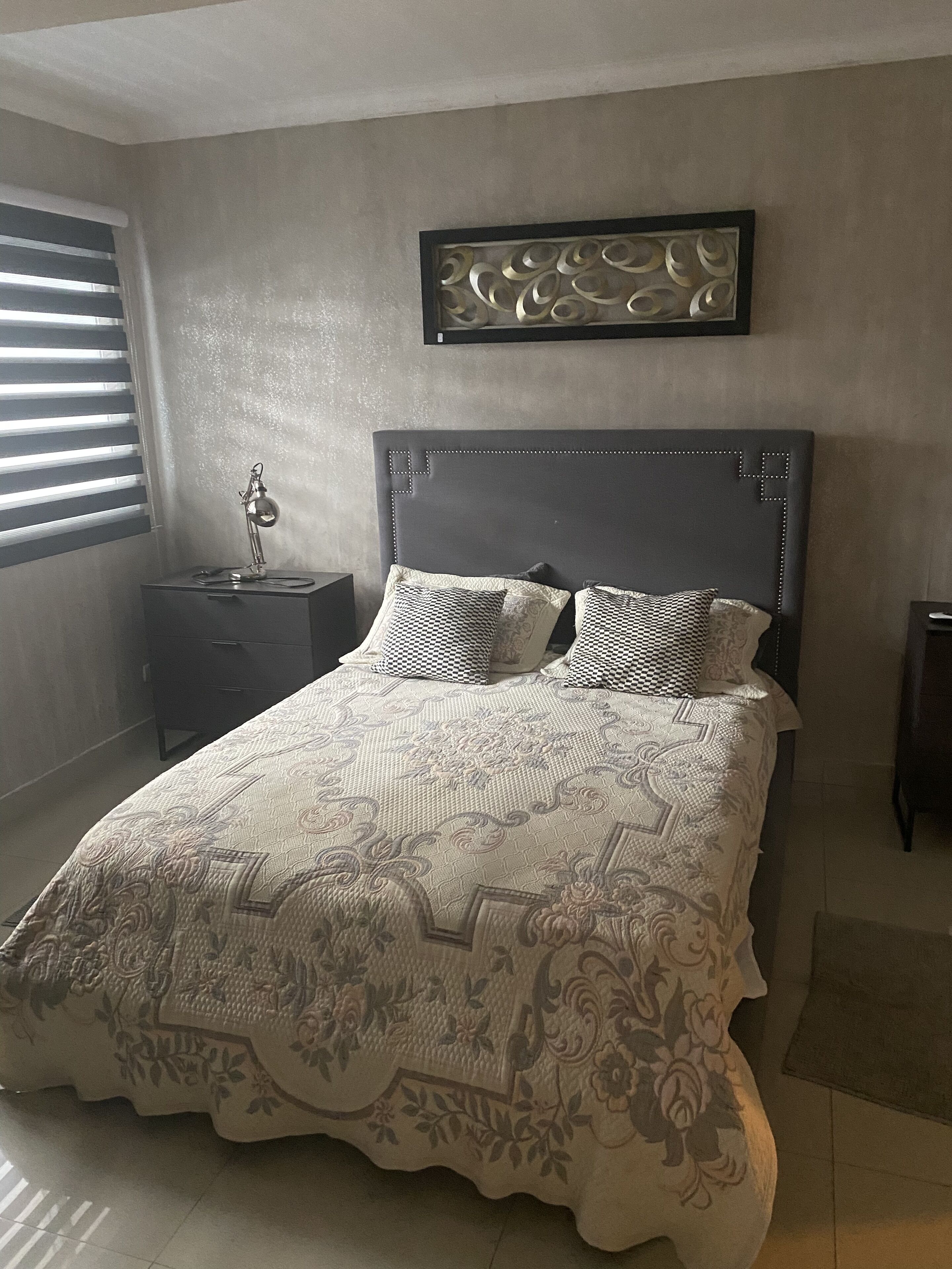 Une chambre à coucher comprenant un lit, une table de chevet, une lampe, une œuvre d’art encadrée accrochée au mur et une fenêtre avec des stores.