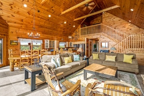 A spacious wooden cabin interior with a living area, dining space, and a staircase leading to an upper level.