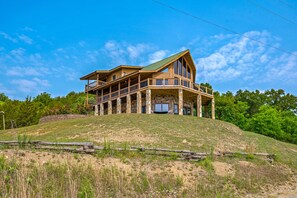 A large, multi-story wooden house with a balcony on the second floor, set on a grassy hill with trees in the background.