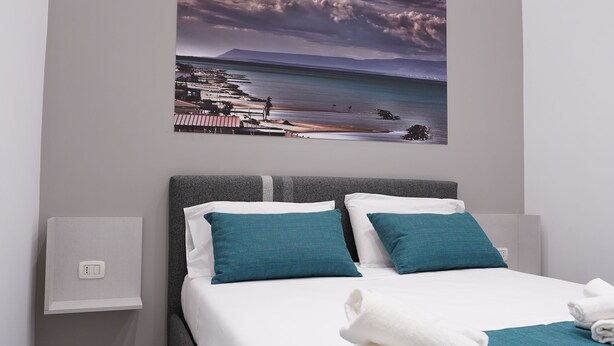 A modern bedroom with a bed, teal bedding, a painting of a coastal scene, and a bedside table. - Puglia in love (Margherita di Savoia)