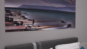 A bedroom with a large landscape painting of a coastal scene, a bed with white and teal pillows, and a gray headboard. - Puglia in love (Margherita di Savoia)