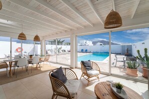 Une terrasse ensoleillée, meublée de mobilier en osier, avec une piscine et une vue sur des bâtiments blancs et des montagnes.