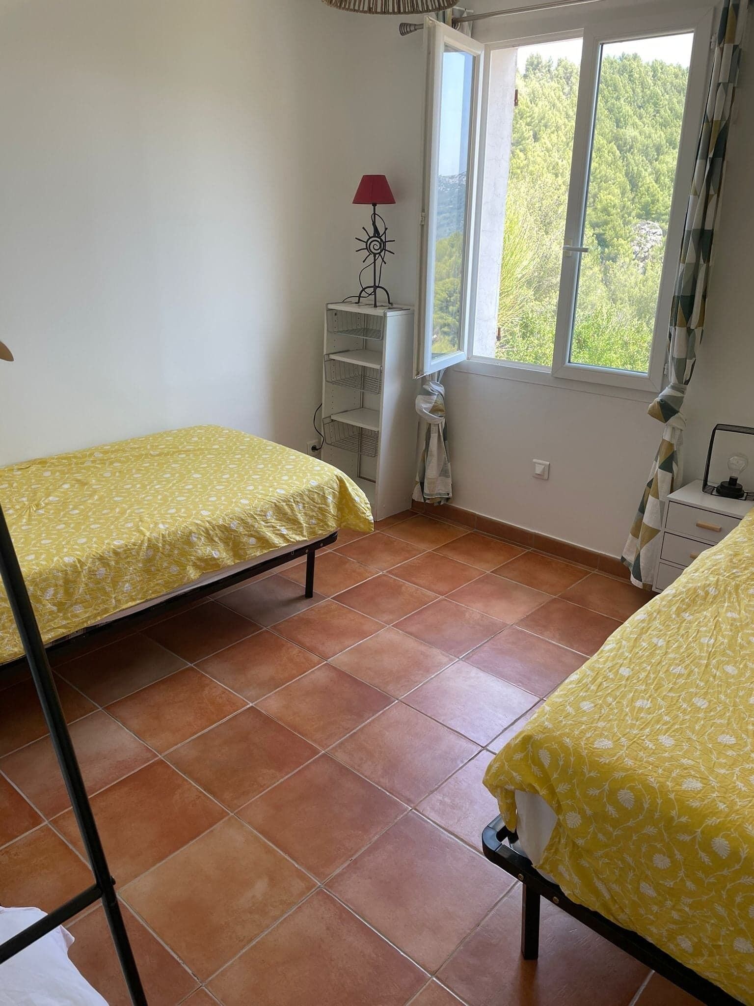 Una habitación con dos camas individuales, una ventana con vistas a zonas verdes, un estante blanco con una lámpara y un suelo de baldosas.