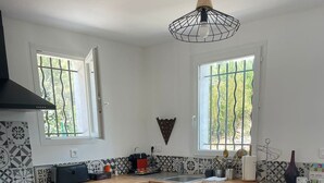 A compact kitchen with a stove, oven, and dishwasher. It features a window with a view of greenery outside, a hanging light fixture, and a tiled backsplash. - T5 house overlooking Toulon harbour with swimming pool (Le Revest-les-Eaux)