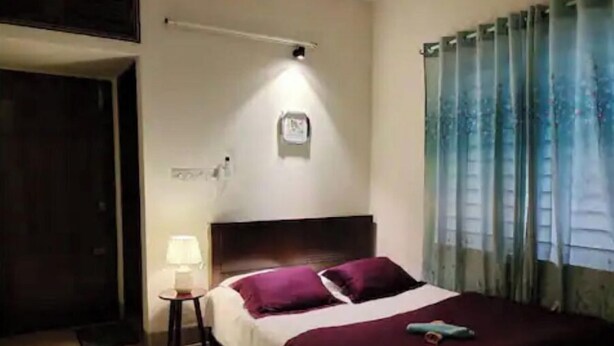 A bedroom with a bed, a nightstand, a lamp, a window with curtains, and a wall-mounted light fixture. - 3 Bedroom Apartment near Dhanmondi & Panthpath (Dhaka)