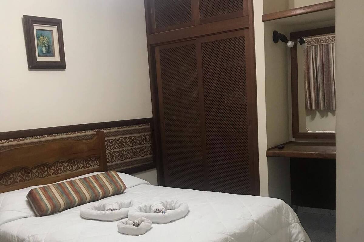 A neatly made bed with a striped pillow, a headboard with a decorative pattern, a wooden wardrobe, and a framed picture on the wall.