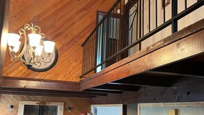 Amplia sala de estar con techo alto, vigas de madera, una araña, chimenea, sofá con una manta de Yellowstone National Park, televisión y un balcón con barandilla.