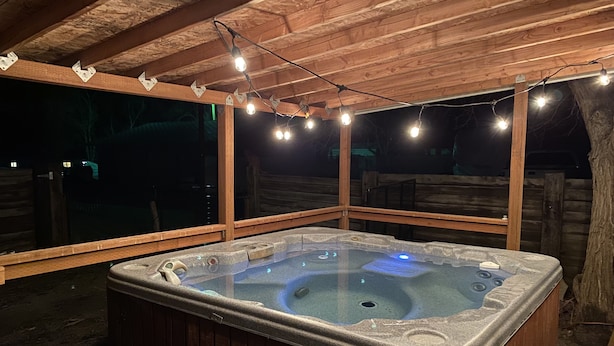 Un jacuzzi bajo una pérgola de madera con luces de cadena, rodeado por una terraza de madera y una valla de madera.